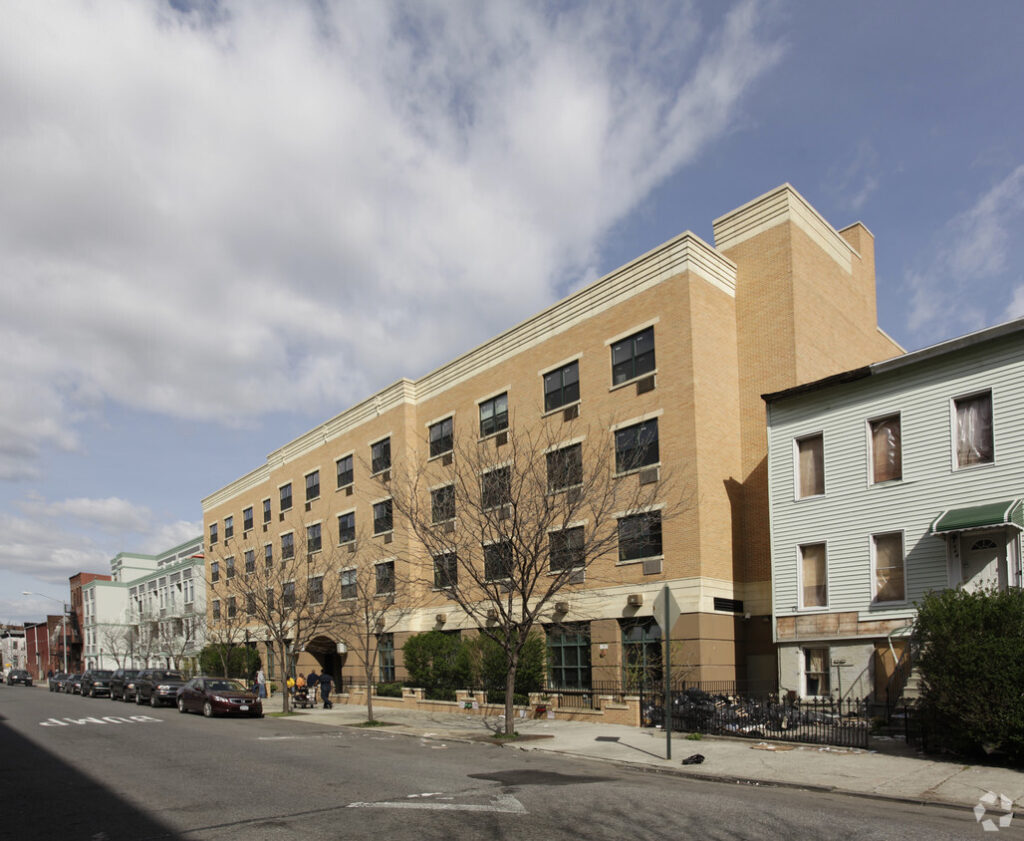 Clover Hall, a modern brick apartment building with dark-framed windows, stands on a city street lined with parked cars and bare trees under a partly cloudy sky.
