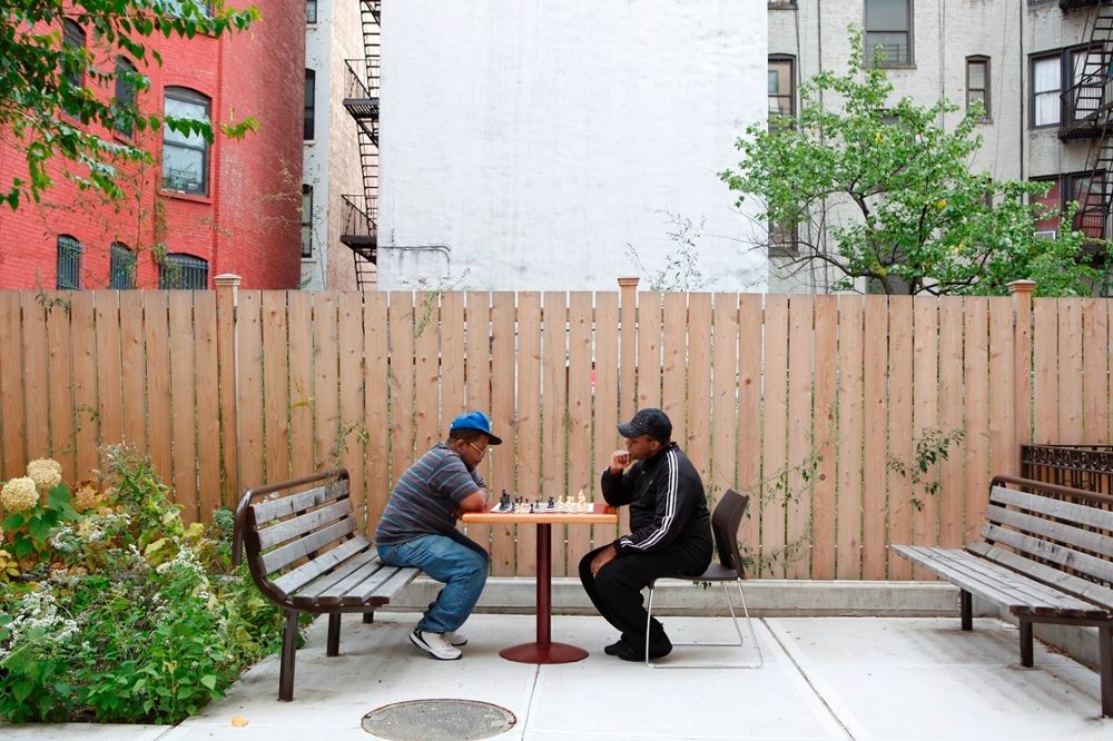 Two men are engaged in a game of chess at a small round table in an outdoor urban setting, flanked by wooden benches and greenery, with brick apartment buildings forming the backdrop.